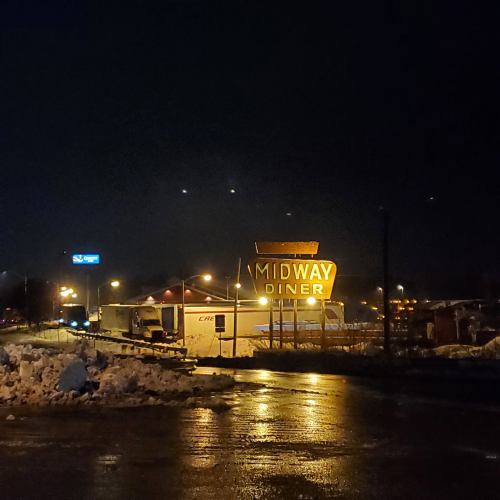 A diner sign illuminated at night, surrounded by snow and wet pavement, with a truck parked nearby.