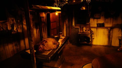 A dimly lit room with a bloodied figure lying on a table, surrounded by eerie decor and shadows.