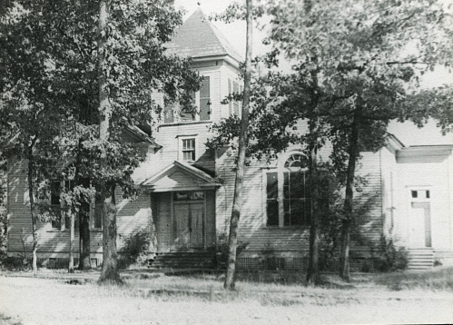 Black and white photo of a large, old house surrounded by trees, featuring a prominent entrance and multiple windows.
