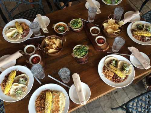 A table set with multiple plates of food, including tacos, corn, rice, beans, chips, and various dips, surrounded by drinks.