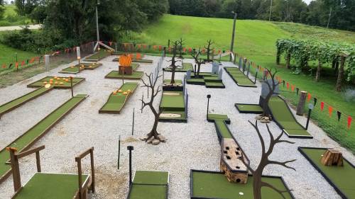 Aerial view of a mini-golf course featuring themed obstacles, trees, and a pumpkin, set in a grassy area.