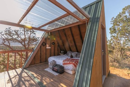 Cozy A-frame cabin with a bed, wooden deck, and large windows, surrounded by trees and natural scenery.