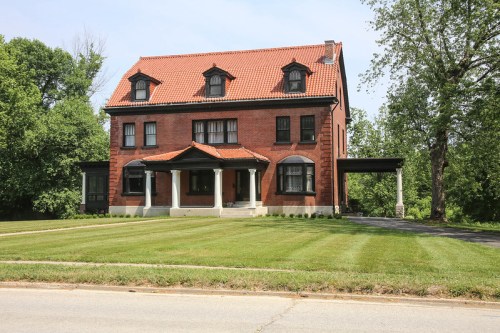 A large red brick house with a tiled roof, featuring a front porch and surrounded by a well-maintained lawn and trees.