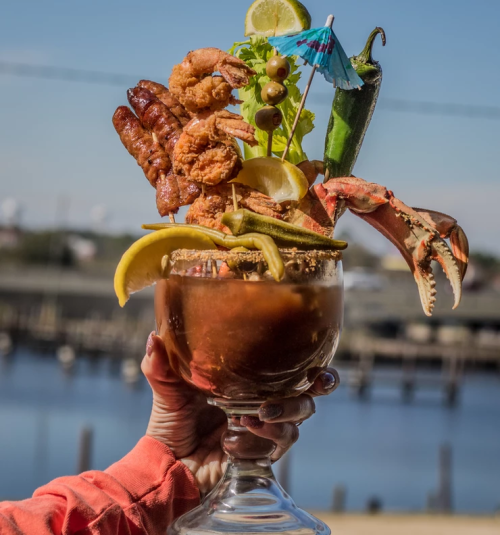 A hand holds a large seafood cocktail with shrimp, crab, vegetables, and garnishes, set against a waterfront background.