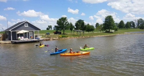 Four people kayaking on a calm lake, with a house and green trees in the background under a blue sky.