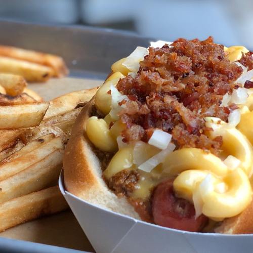 A hot dog topped with macaroni and cheese and crispy bacon bits, served with a side of golden fries.