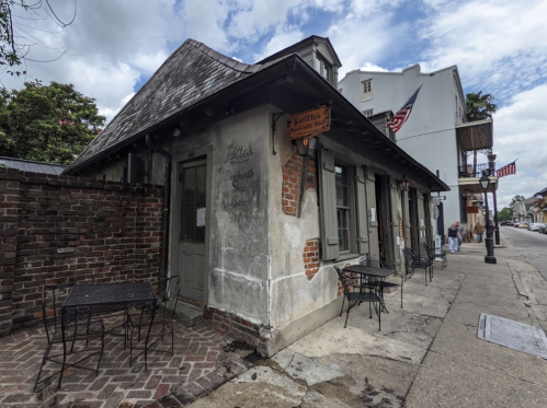 Historic building with a weathered facade, outdoor tables, and a sign, set on a charming street under a cloudy sky.