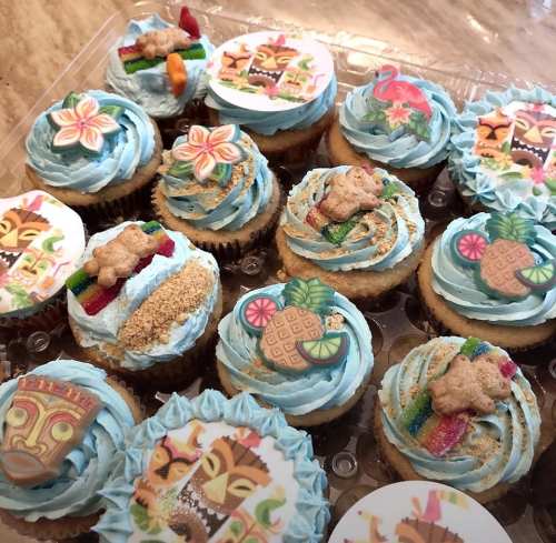 A tray of colorful cupcakes with blue frosting, tropical decorations, and themed toppings like tiki faces and pineapples.