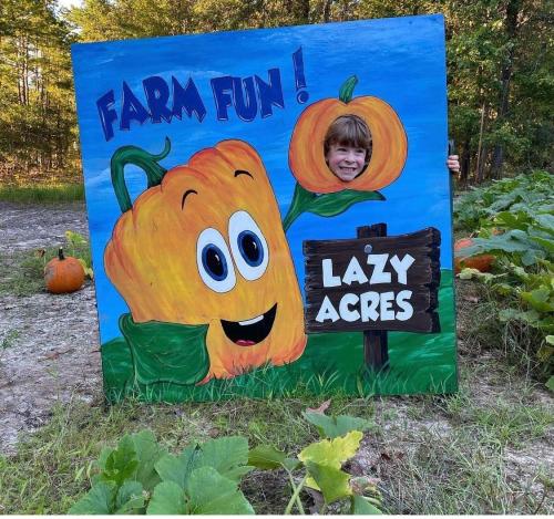 A colorful farm-themed sign with a smiling bell pepper character and a girl peeking through a cutout at Lazy Acres.