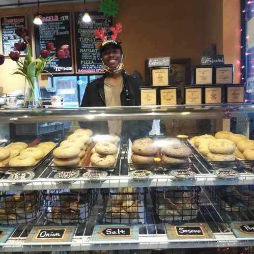A smiling person stands behind a display case filled with various bagels in a cozy café setting.