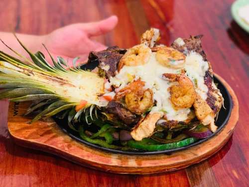 A sizzling dish featuring grilled shrimp and chicken served on a pineapple, with melted cheese and vegetables.