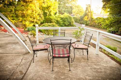 A patio with a round table and four chairs, surrounded by greenery and a quiet street in the background.