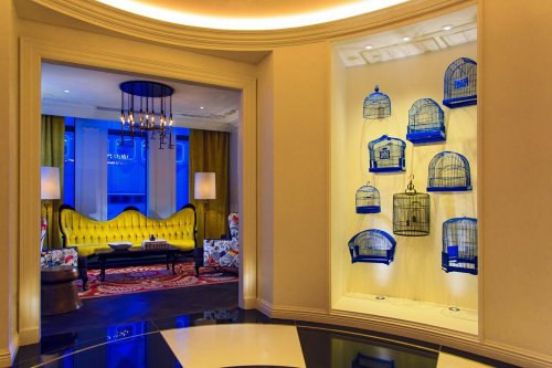 A stylish interior featuring a yellow sofa, patterned chairs, and decorative birdcages on a wall. Soft blue lighting enhances the ambiance.