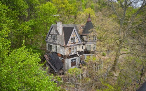 An abandoned, dilapidated house surrounded by lush green trees and overgrown vegetation.