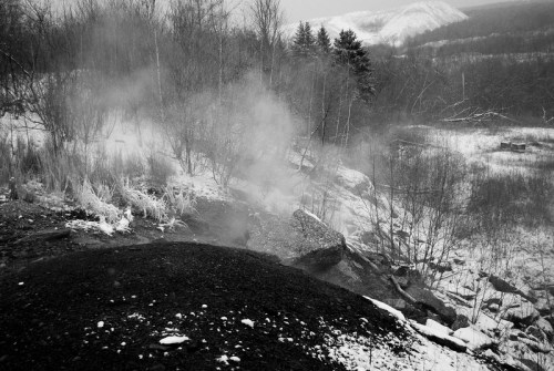 A misty, snowy landscape with steam rising from a rocky area, surrounded by bare trees and distant hills.
