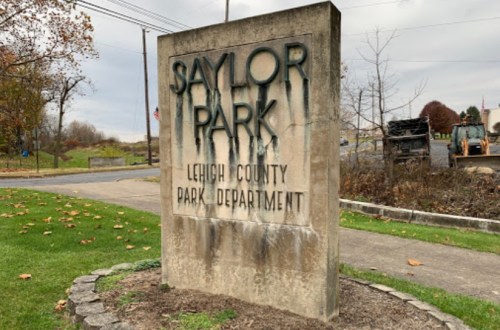 Sign for Saylor Park, Lehigh County Park Department, with weathered appearance and surrounding grass and trees.