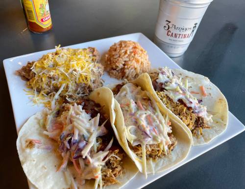 Three tacos filled with shredded meat and slaw, served with rice, beans topped with cheese, and a drink on the side.