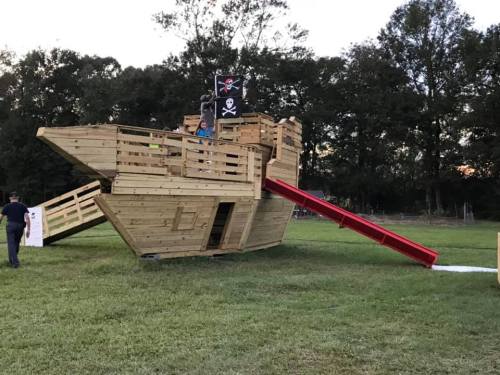 A wooden pirate ship play structure with a slide, set in a grassy area surrounded by trees.