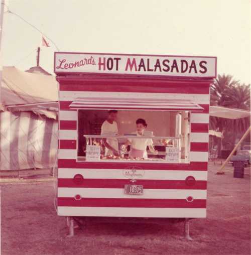 A vintage food truck with red and white stripes, featuring "Leonard's Hot Malasadas" sign and two people inside.