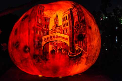 A carved pumpkin glowing orange, depicting a scenic canal with buildings and a gondola at night.