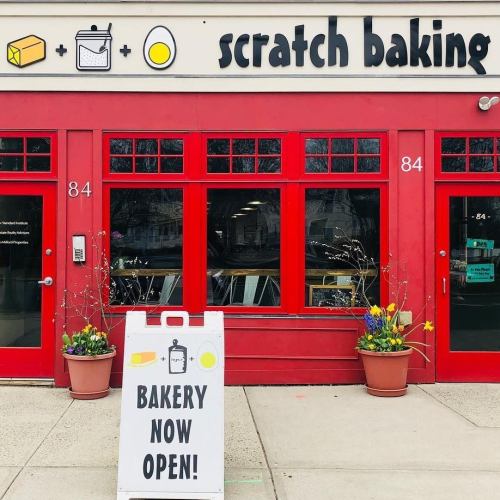 Exterior of Scratch Baking bakery with red doors, a sign reading "Bakery Now Open," and potted plants on either side.