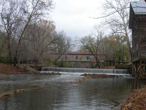 A serene river scene featuring a small waterfall, surrounded by trees and an old wooden building in the background.