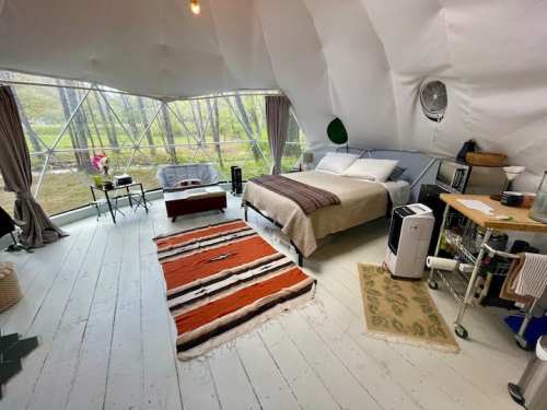 Cozy, modern interior of a geodesic dome with a bed, seating area, and large windows overlooking nature.