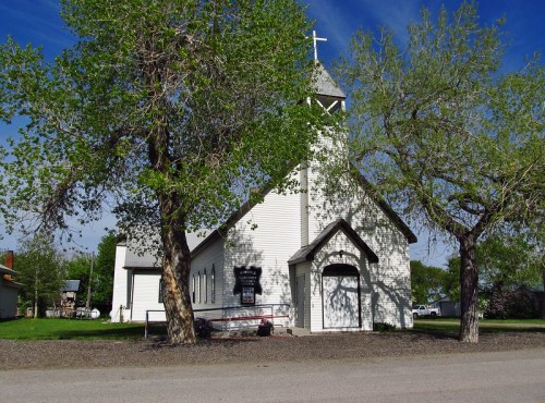 A white church with a steeple, surrounded by green trees and a clear blue sky.