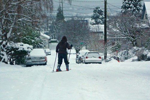 A person skiing down a snowy street lined with parked cars and trees, with a winter landscape in the background.