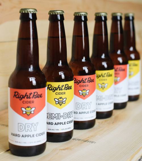Six bottles of Right Bee Cider in varying labels: Dry, Semi-Dry, and Dry Hard Apple Cider, arranged on a wooden surface.