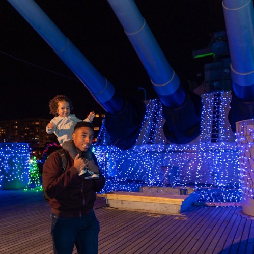 A man carries a girl on his shoulders, surrounded by colorful lights on a festive night.