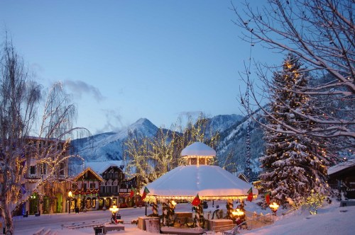 A snowy village scene at dusk, featuring a lit gazebo, festive decorations, and mountains in the background.