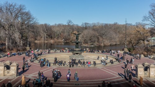 A busy park scene featuring a large fountain surrounded by people, with trees and a lake in the background.