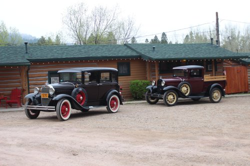 Two vintage cars parked in front of a log cabin, surrounded by trees and a gravel driveway.