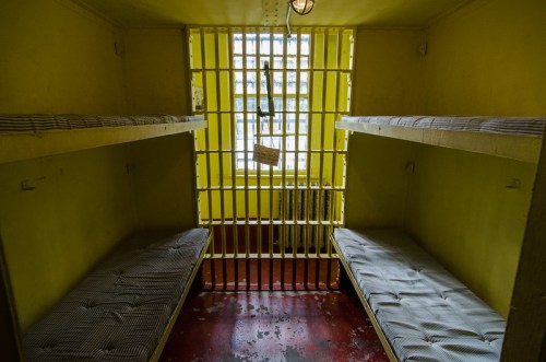 Interior of a small, yellow prison cell with two bunk beds and barred windows.