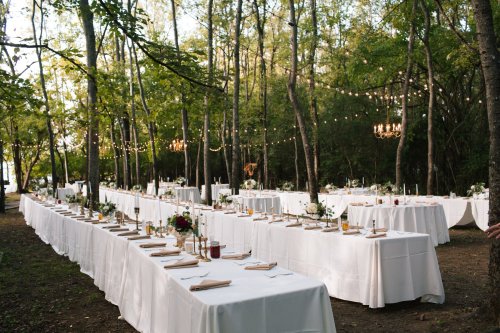 A beautifully arranged outdoor dining setup with white tables, floral centerpieces, and string lights in a forest setting.