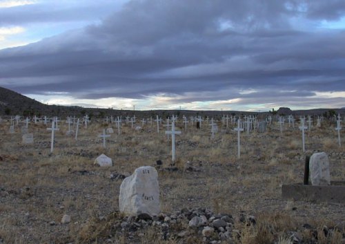 A vast cemetery with numerous white crosses and a cloudy sky overhead, set in a dry, rocky landscape.