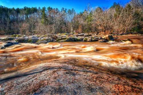 A flowing river with brown water over rocky terrain, surrounded by trees under a clear blue sky.