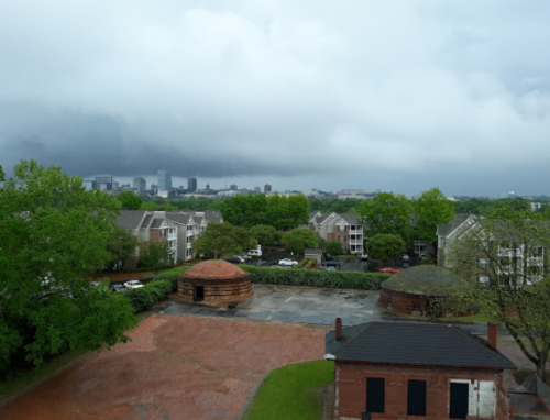 A cloudy sky looms over a residential area with green trees and historic buildings in the foreground.