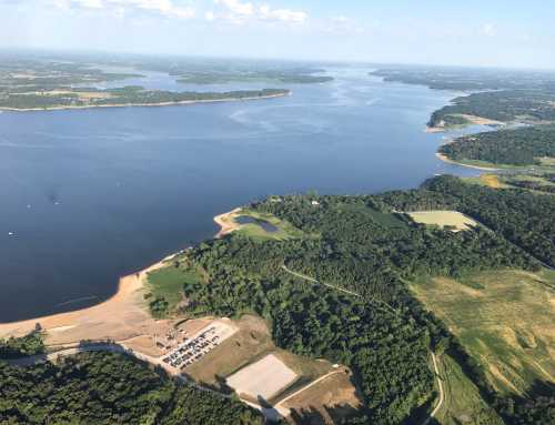 Aerial view of a large lake surrounded by green forests and fields, with a sandy beach and parking area visible.