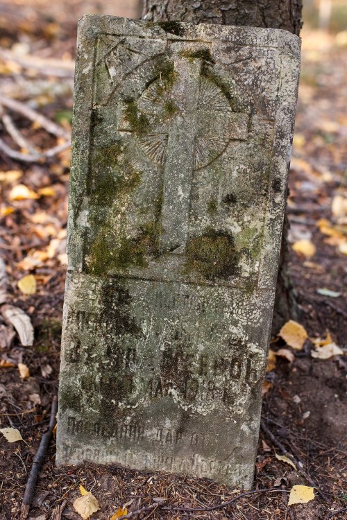 A weathered gravestone with a cross design, partially covered in moss, set in a forested area with fallen leaves.