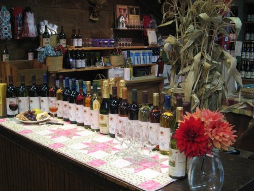 A cozy wine shop display featuring various bottles of wine, glasses, and decorative flowers on a quilted tablecloth.
