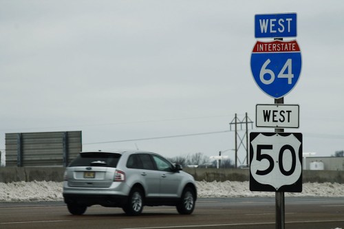 A silver SUV drives past a highway sign for Interstate 64 West and U.S. Route 50 West on a cloudy day.