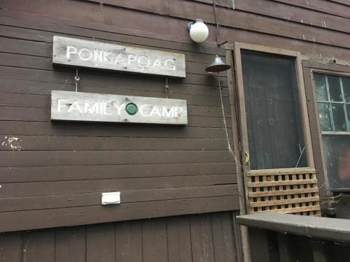 Wooden signs reading "Ponkapoag Family Camp" on a rustic cabin wall, with a door and window nearby.