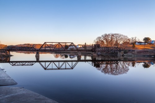 A tranquil scene of a bridge reflected in calm water, surrounded by trees and a clear blue sky at sunset.