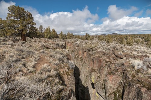 A rocky landscape with a deep crevice, surrounded by sparse vegetation and trees under a partly cloudy sky.