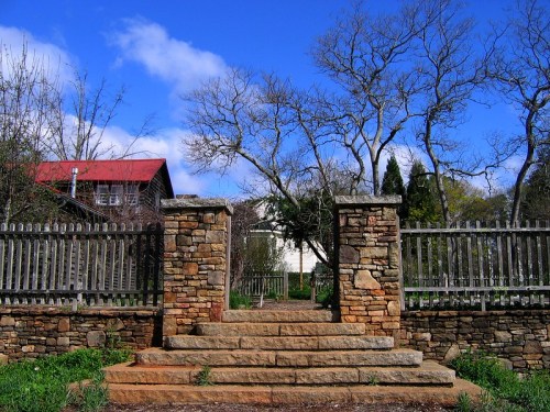 Stone steps lead through a rustic gate, framed by trees and a blue sky, with a house visible in the background.