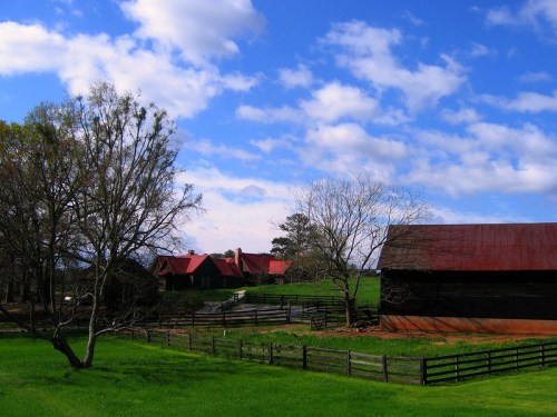 A scenic farm landscape featuring a red-roofed barn, a house, and green fields under a blue sky with clouds.