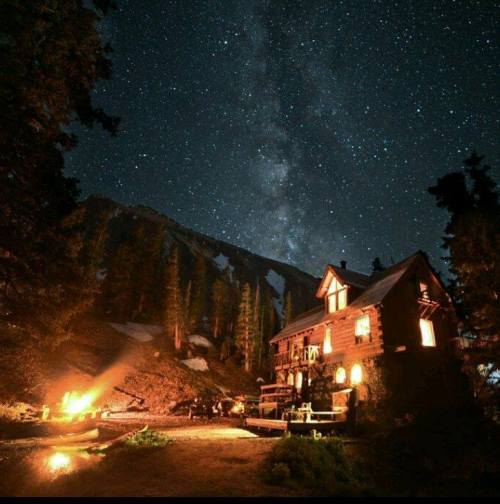 A cozy cabin under a starry sky, with a campfire nearby and mountains in the background.