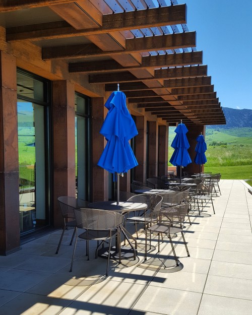 A sunny patio with blue umbrellas and tables, surrounded by green fields and mountains in the background.